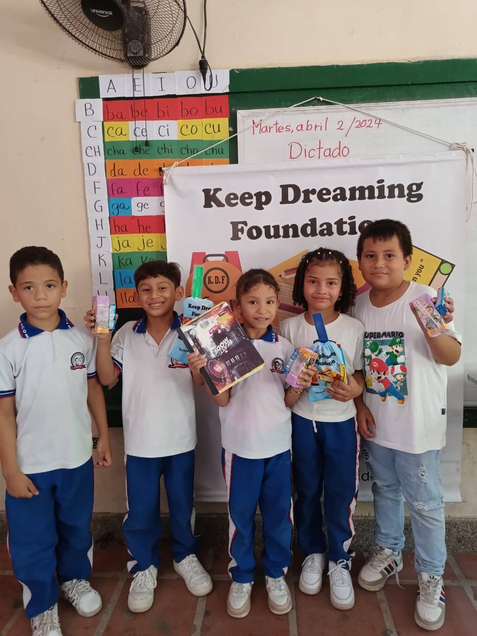 Five children holding KDF kits in a classroom, with a Spanish phonics chart and the date Martes abril 2/2024 on the board behind them